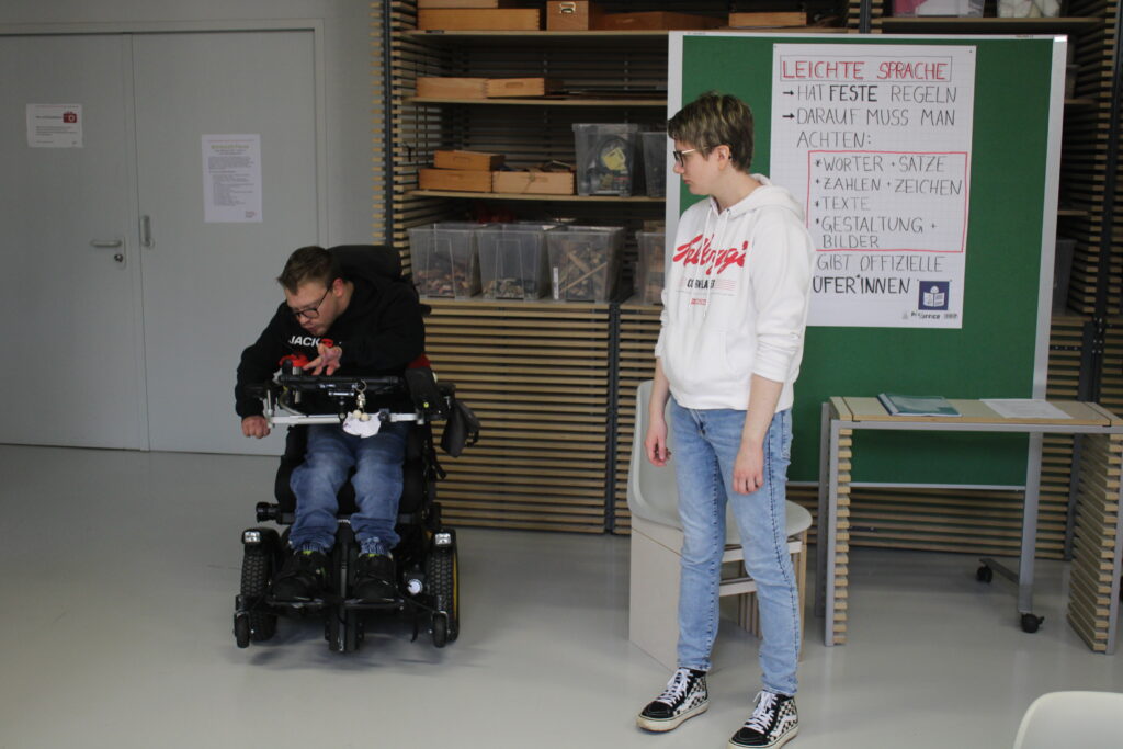 Zwei Bildungsfachkräfte aus dem Forum für inklusive Bildung. Einer der beiden Personen sitzt im Rollstuhl und berichtet uns wichtige Dinge über barriereärmere Sprache und Umgang für Menschen mit Behinderung über seinen Sprachcomputer.